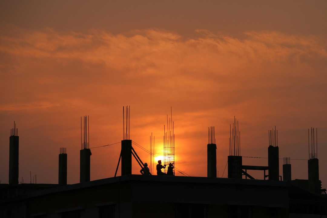Construction workers silhouetted against an orange sunset on a high-rise building site with exposed rebar columns